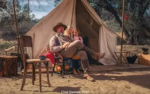 A family sitting outside a canvas tent in a rural setting.