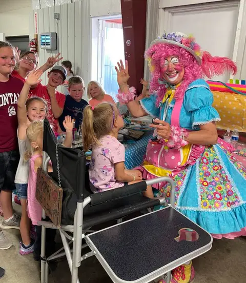 A clown in colorful costume interacts with children indoors.