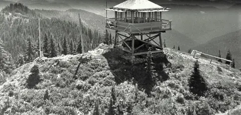 A fire lookout tower sits atop a mountain peak overlooking a forested landscape.