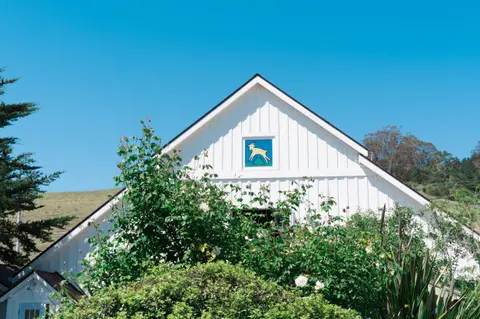 A white barn with a goat logo sits among greenery under a clear blue sky.