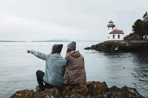 Two people sit on rocks by the water and look at a distant lighthouse.