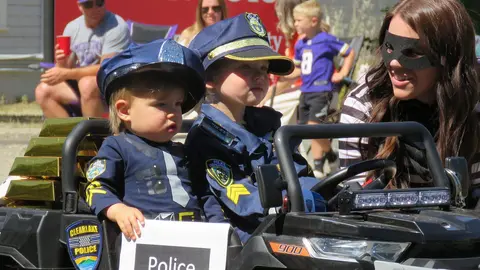 Children dressed as police officers ride in a toy vehicle alongside someone in a burglar costume.
