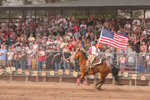 A person on a horse carries an American flag in an arena before a crowd.