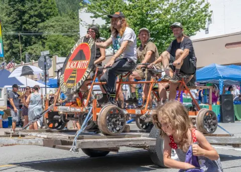 Several people ride a large multi-person bicycle on a trailer during an outdoor event.