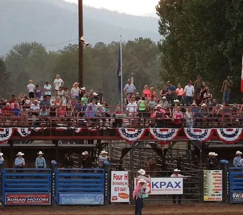 A crowd watches an event from stands decorated with patriotic bunting at a rodeo arena.