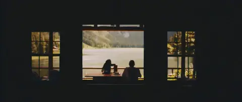 People sit at a table overlooking a lake through a window.