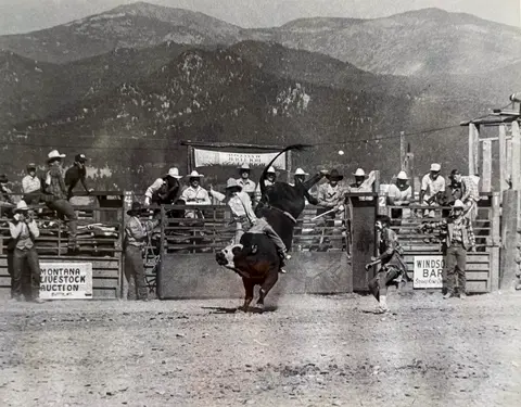 A historic black and white photo shows a bull rider at a rodeo event with mountains in the background.