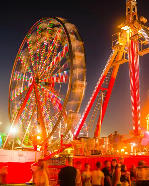 A brightly lit Ferris wheel and carnival ride at night with people gathered in the foreground.