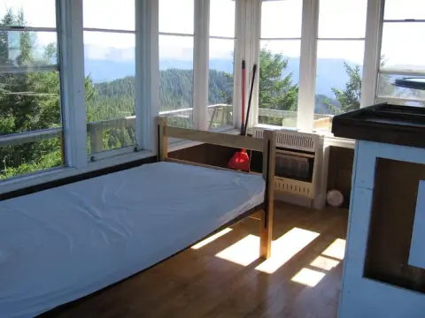 A single bed inside a room with large windows overlooking a forest-covered mountain range.