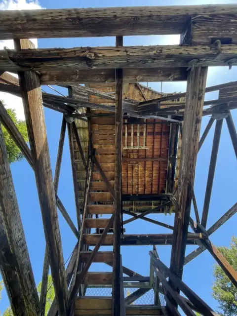 A low-angle view looking up at the wooden structure and stairs of a tower against a blue sky.