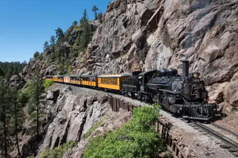 A steam train travels along a narrow cliffside track.