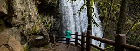 A person stands on a trail looking at a large waterfall.