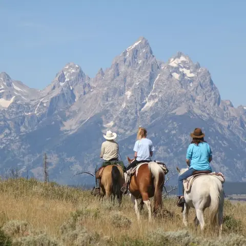 Three people on horseback riding through a grassy field with mountains in the background.