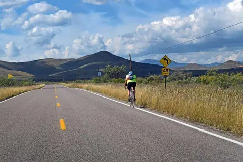 A cyclist rides along the edge of a paved road beneath a cloudy sky and distant hills.