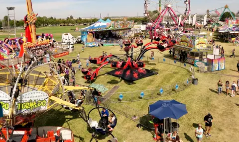 An aerial view shows an outdoor carnival with various rides, food stands, and visitors walking on grassy grounds.