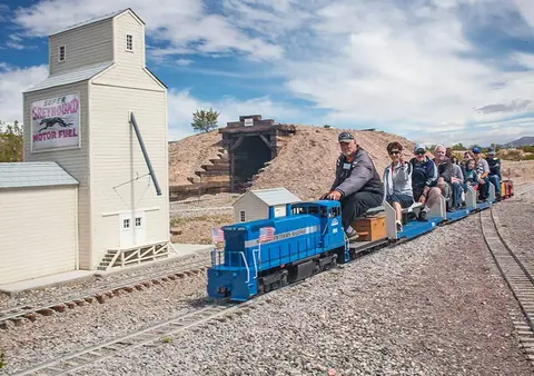 People ride a small scale model train past miniature buildings.