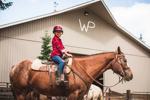 A child wearing a helmet and a red plaid shirt sits on a brown horse outside a barn.