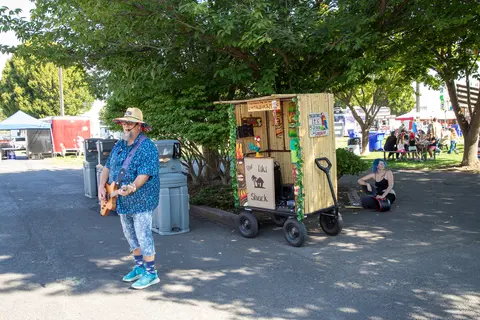 A man plays a guitar in front of a small tiki shack cart outdoors.