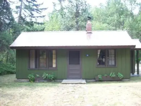 A single-story green cabin with a white roof is surrounded by trees.