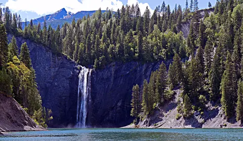 A tall waterfall cascading down a cliff into a lake surrounded by evergreen trees and mountains.
