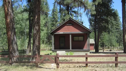 A red log cabin stands among tall pine trees behind a wooden fence.