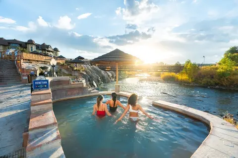 Three people relax in an outdoor hot spring pool near a river at sunset.