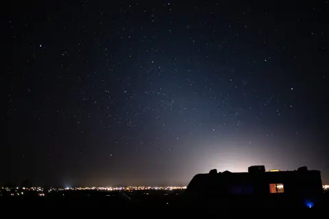 A starry night sky over a silhouette of a vehicle and distant city lights.