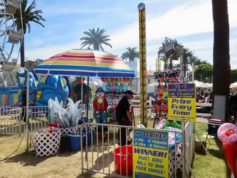 A carnival game booth with a striped umbrella stands outdoors near palm trees and amusement rides.