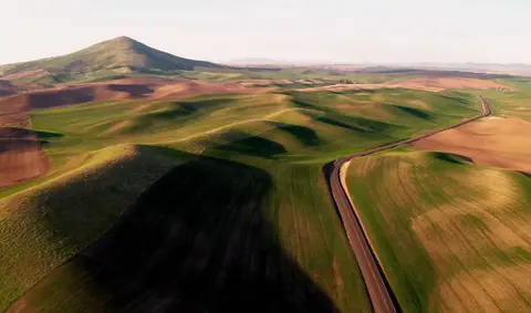 An aerial view of a winding road through rolling green and brown hills with a large hill in the background.
