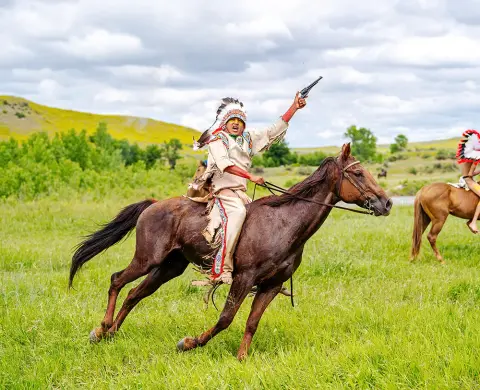 A person in traditional indigenous attire rides a horse across a grassy field, holding a pistol in the air.