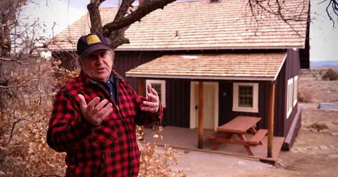 A man in a red flannel shirt stands outside a dark wooden cabin with a shingled roof.