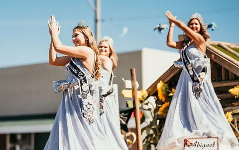 Three young women in gowns and tiaras wave from a parade float.