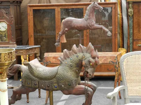 An outdoor antique market display features two wooden carousel horses and other antique furniture.