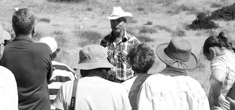 A group of people listening to a man wearing a cowboy hat outdoors.