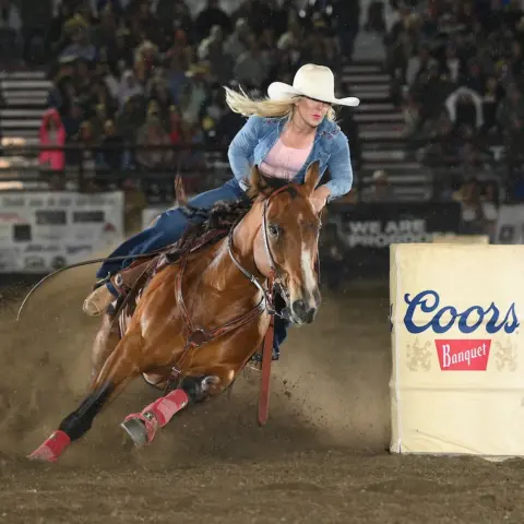 A cowgirl rides a horse sharply turning around a barrel in an arena.