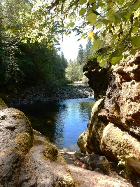 A river flows between mossy rocks and trees under a bright sky.