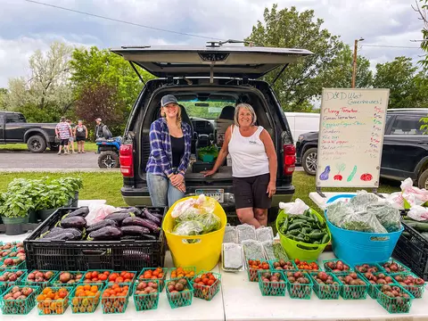 Two women pose behind a vegetable stand selling produce out of the back of an SUV.