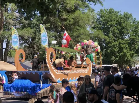 A parade float designed like a boat with people on it, surrounded by a crowd.