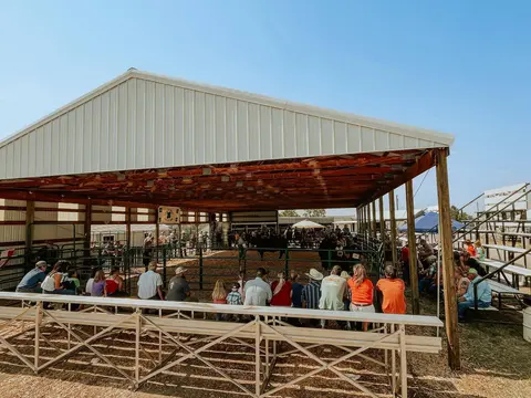 A crowd of people sitting and watching an event inside an open-sided agricultural pavilion.