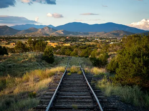 A view of railroad tracks running through a grassy landscape towards mountains under a cloudy sky.