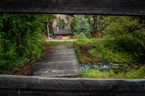 A wooden bridge leads across a stream toward a small log cabin in a wooded area.