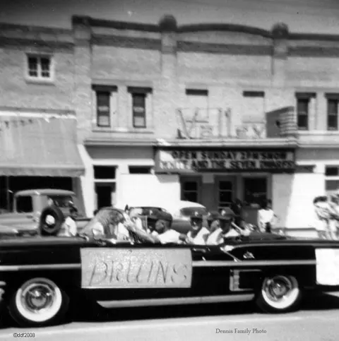 Black and white historic photo showing people in a convertible car driving past a building.