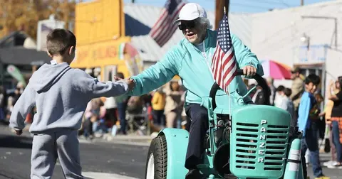An older adult on a small teal tractor reaches out to high-five a young boy.