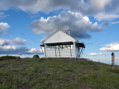 A white cabin sits atop a grassy hill under a cloudy blue sky.