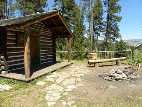 A log cabin sits in a forest with a stone path and fire pit outside.