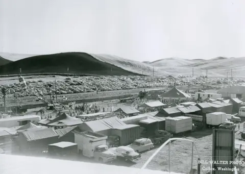 A black and white photograph of a fairgrounds with tents and vehicles set against rolling hills.