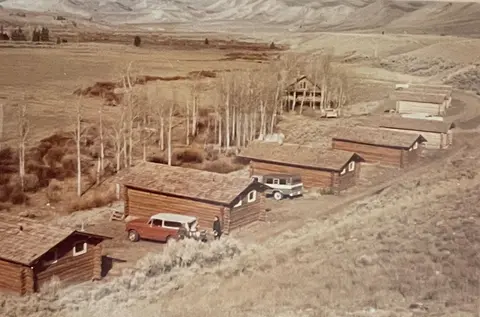 A high-angle view shows a row of log cabins in a dry landscape with mountains in the background.