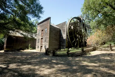 A historic wooden mill with a large water wheel surrounded by trees and a rustic fence.