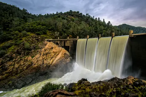 Water spills over a dam surrounded by trees and rocky hills.