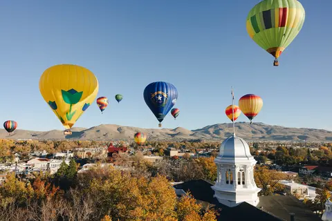 Hot air balloons fly over a town with autumn trees and mountains in the distance.